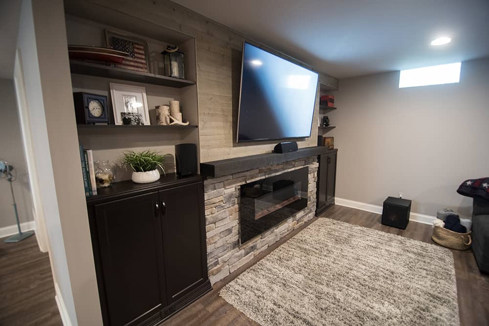 Tv wall in basement with wood beam and electric fireplace wrapped in stone in Wixom, Michigan