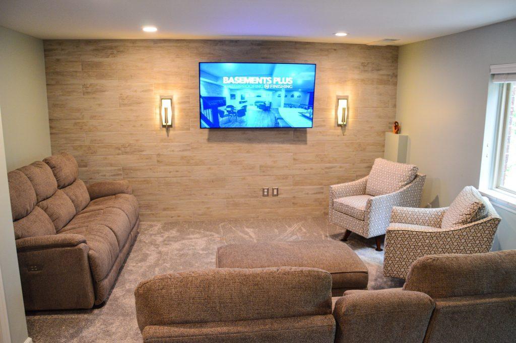 Finished basement living room area with two couches and chairs for seating, light tan color shiplap and decorative lights on each side of the tv.