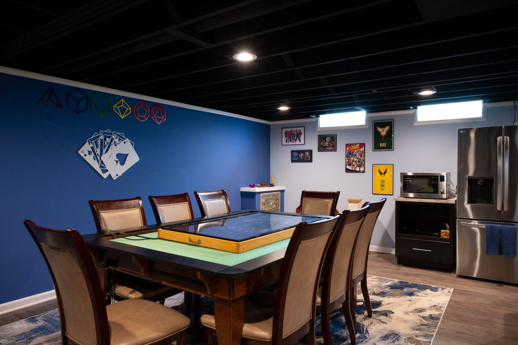 Finished basement "man-cave" with wood floor. A wood game table surrounded by chairs sits in the center. A blue accent wall with various color game dice and playing cards adorn the wall. In the back, a microwave and stainless steel fridge. Southfield, Michigan. Remodeled by Basements Plus.
