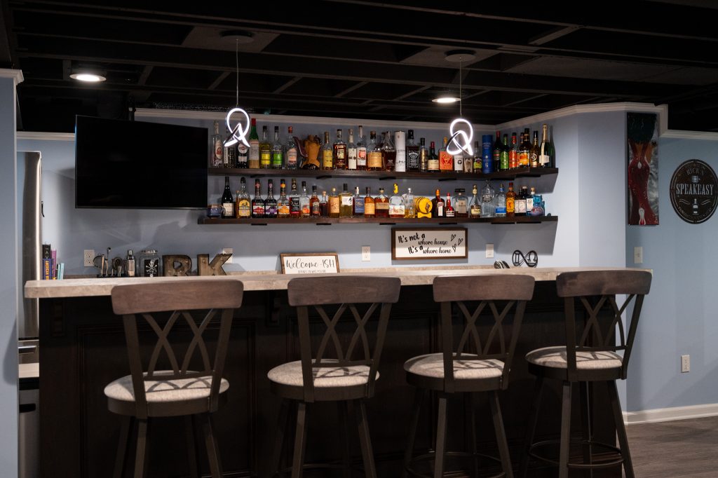 Finished basement "man-cave" with wood floor. A large, angled white couch sits in front of a large screen TV mounted on a dark blue accent recess. A bar with modern hanging lights and wooden barstools to the left. Various bottles of alcohol sit on mounted dark wood shelves. Southfield, Michigan. Remodeled by Basements Plus.