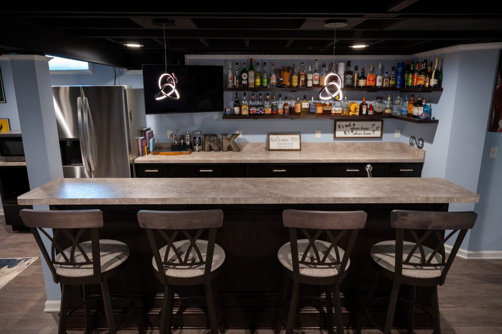 Basement bar with sleek countertop, kitchenette, and shelving with barstools.
