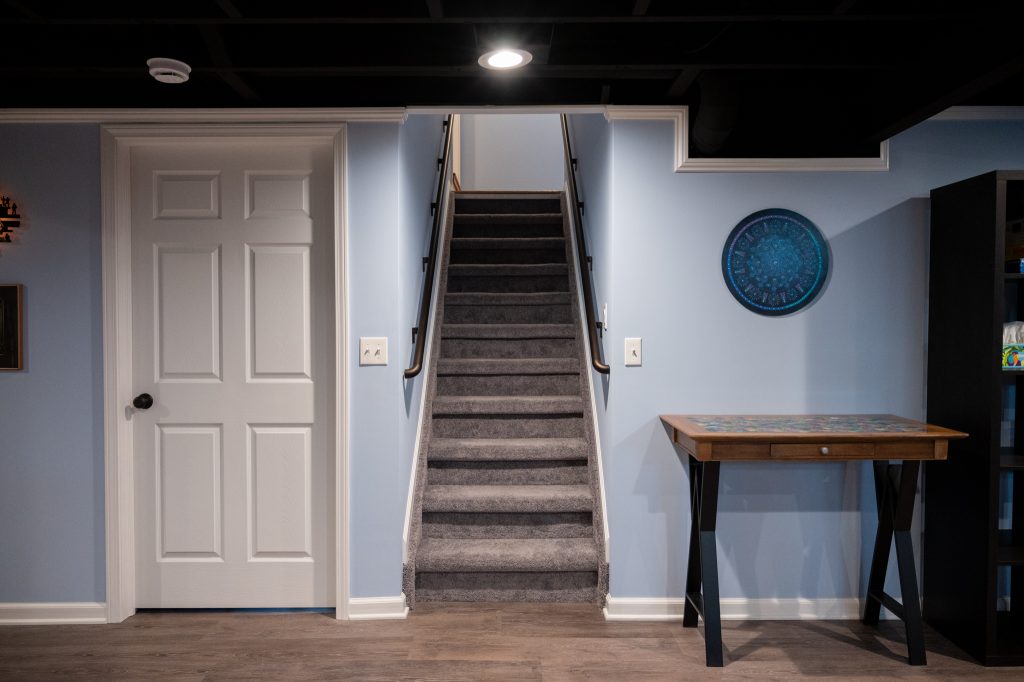 Photo of remodeled basement with grey carpeted stairs, white trim and baseboard, and light blue walls.