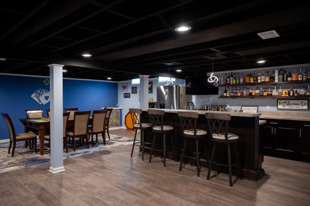 Finished basement "man-cave" with wood floor. A large, angled white couch sits in front of a large screen TV mounted on a dark blue accent recess. A bar with modern hanging lights and wooden barstools to the left. Various bottles of alcohol sit on mounted dark wood shelves. Southfield, Michigan. Remodeled by Basements Plus.