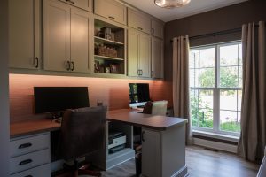 Photo of remodeled home office with dark brown and tan color palette. A Mac computer and monitor on each side of a divider of a built-in desk.